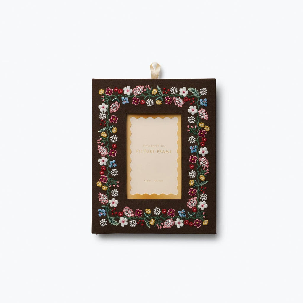 A rectangular dark brown picture frame decorated with a colorful border of small embroidered flowers, hanging from a white ribbon against a light gray background.