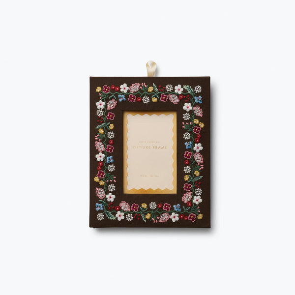 A rectangular dark brown picture frame decorated with a colorful border of small embroidered flowers, hanging from a white ribbon against a light gray background.