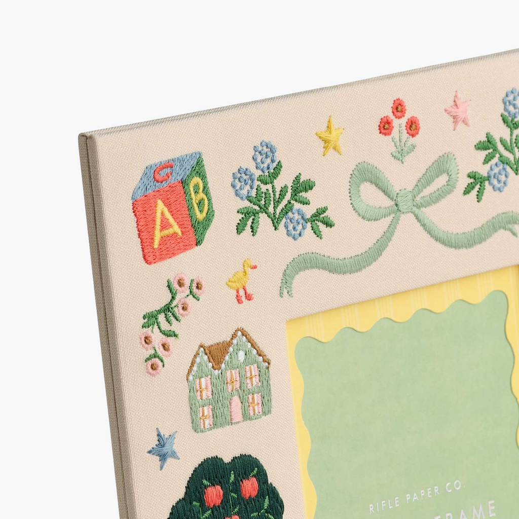 Close-up of a decorative embroidered picture frame corner on a light gray background. The frame has a cream base with colorful floral and nursery-themed embroidery including blue flowers, red berries, yellow stars, a green bow, a small house, and a yellow scalloped mat area.