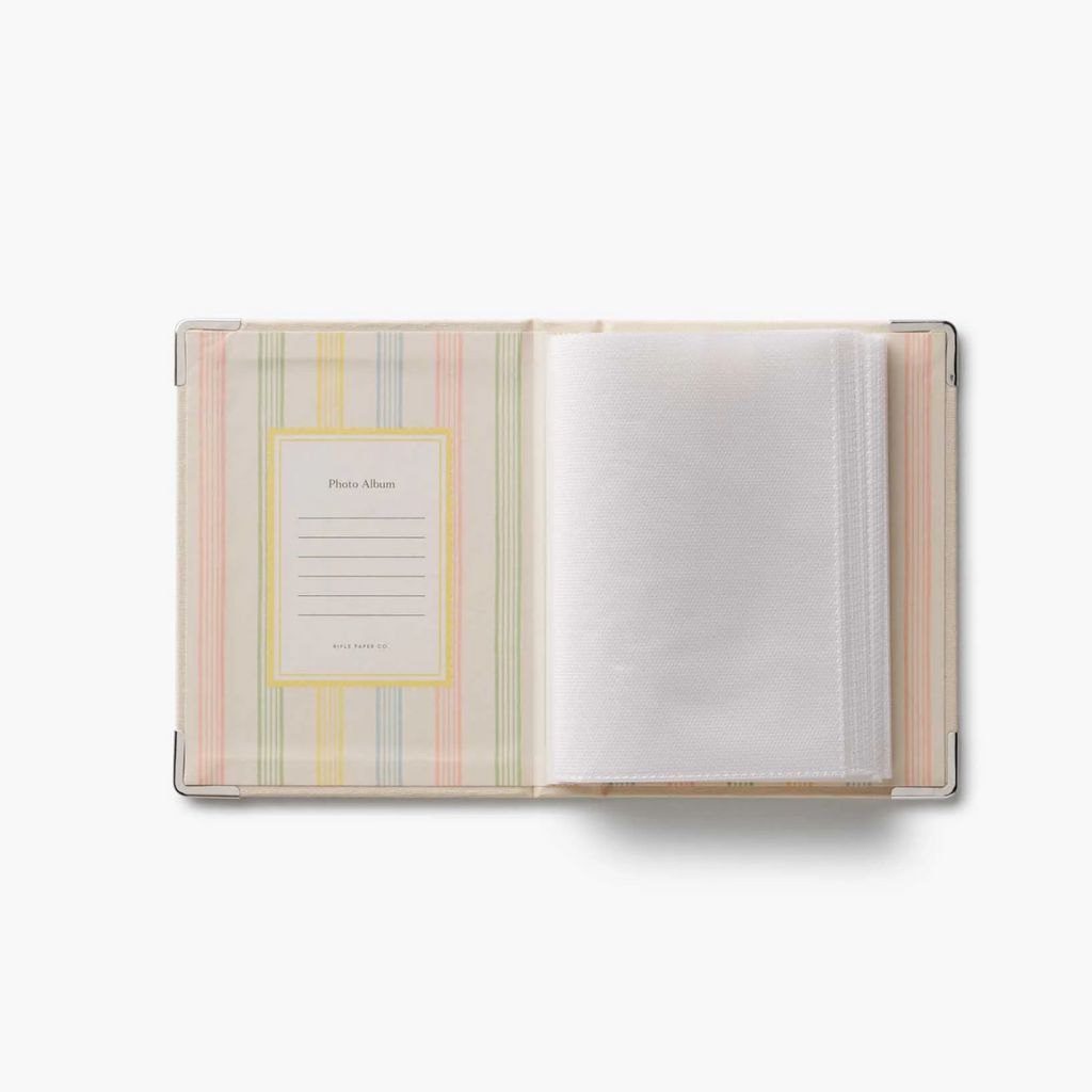 An open photo album with a pastel striped cover in pink, yellow, green, and blue, featuring a photo frame insert on the left and white photo sleeves on the right, set against a plain white background.