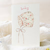 Floral baby bonnet illustration on a textured card with soft focus background