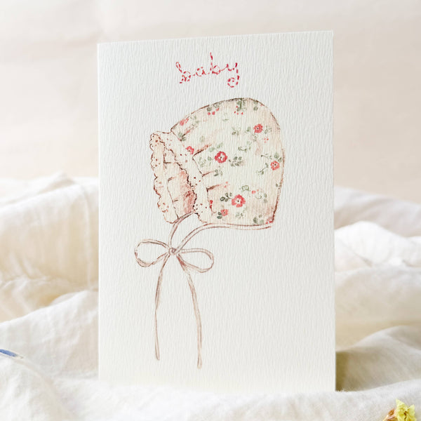 Floral baby bonnet illustration on a textured card with soft focus background