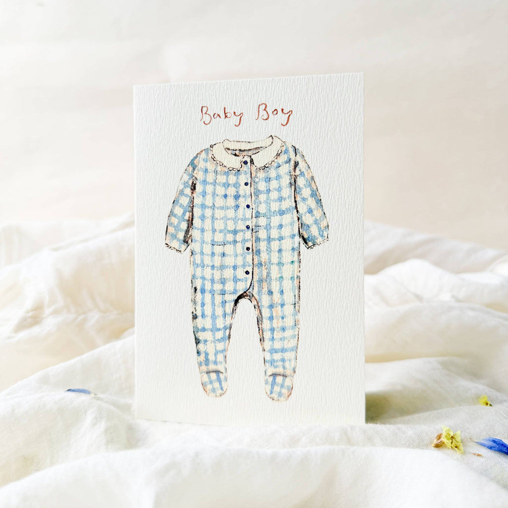 Blue checkered baby onesie on a white card with 'Baby Boy' text, placed on a soft white fabric background.
