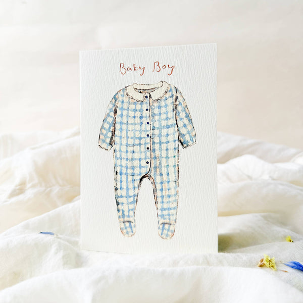 Blue checkered baby onesie on a white card with 'Baby Boy' text, placed on a soft white fabric background.