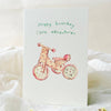 Greeting card with a child's tricycle illustration and 'Happy birthday little adventurer' text on a soft white background.