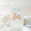 Greeting card with a bicycle illustration and 'Happy birthday little adventurer' text on a white surface.