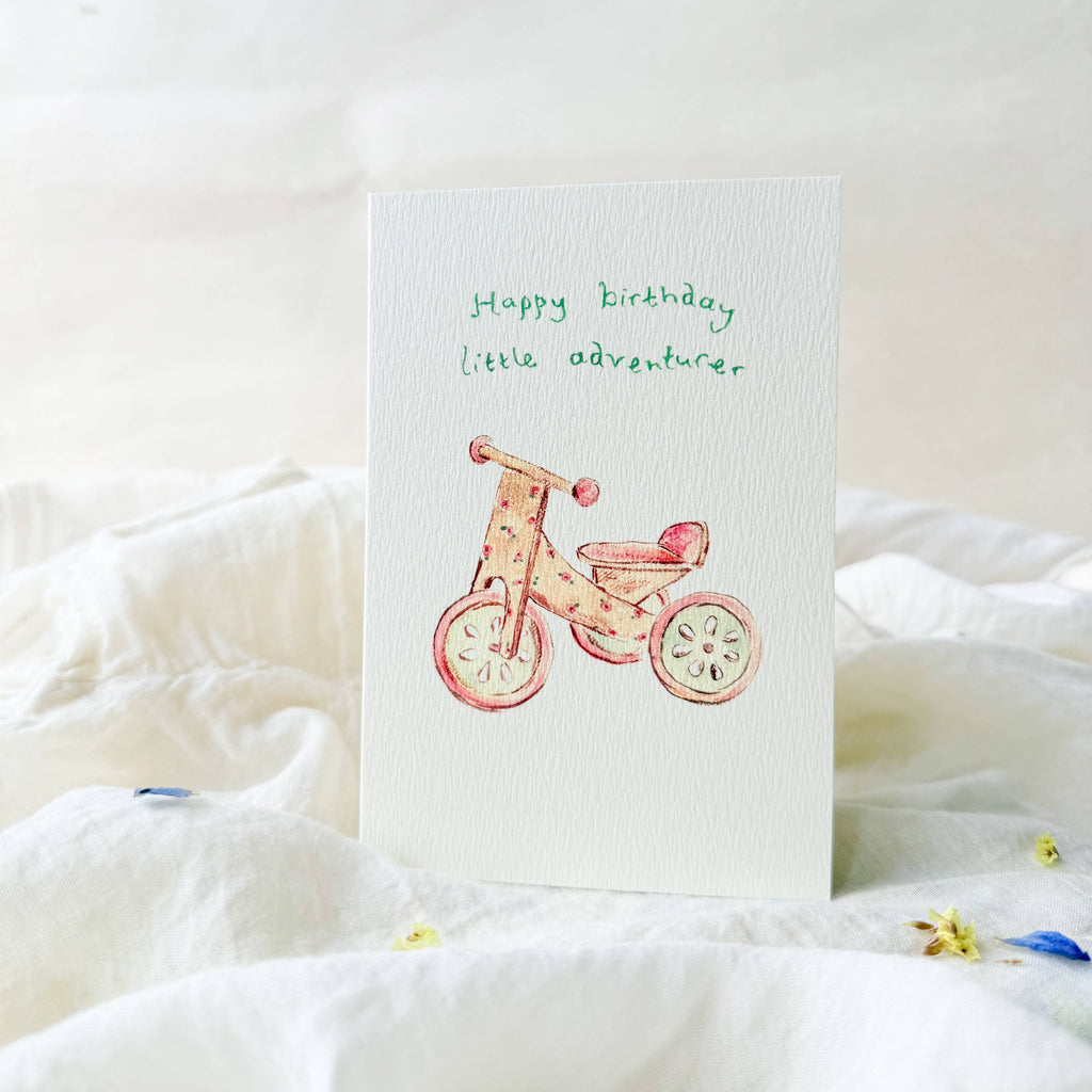 Greeting card with a child's bicycle illustration and 'Happy birthday little adventurer' text on a white surface.