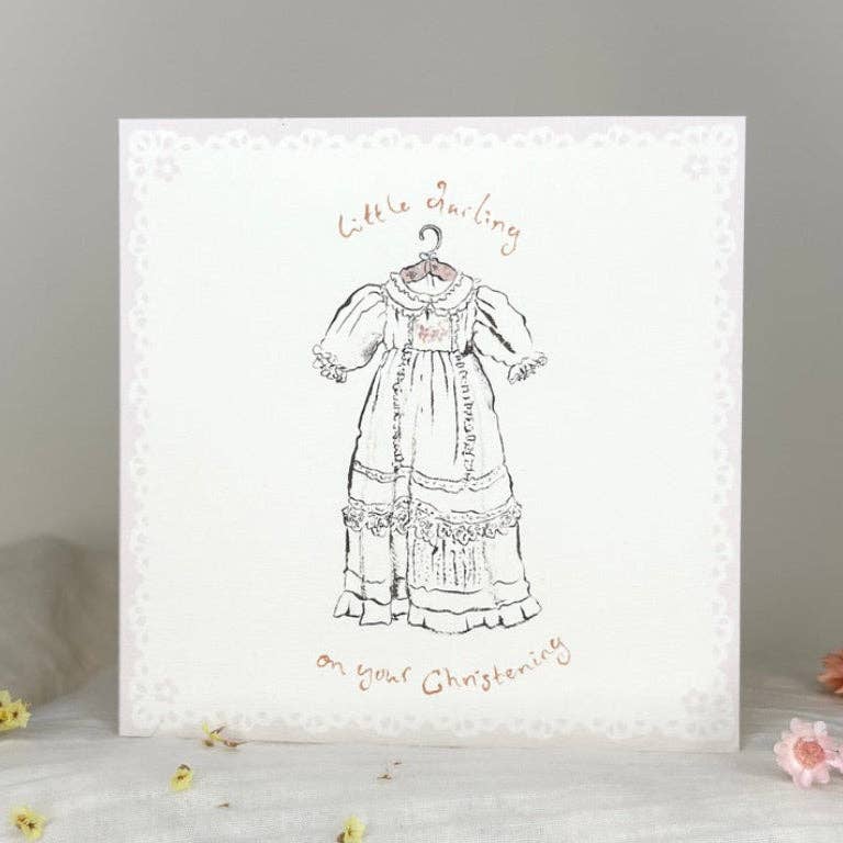 A square white card or print with a delicate lace-like border, featuring a black line drawing of a historical dress on a hanger and the words 'Little Darling on your Christening' in a warm brown script. The item is photographed standing on a soft white surface with small floral props against a light gray background.