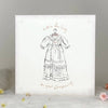 A square white card or print with a delicate lace-like border, featuring a black line drawing of a historical dress on a hanger and the words 'Little Darling on your Christening' in a warm brown script. The item is photographed standing on a soft white surface with small floral props against a light gray background.
