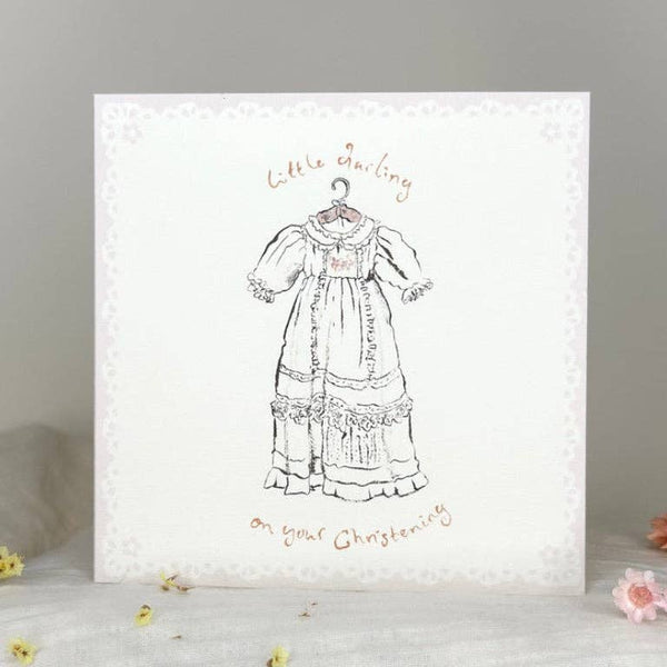 A square white card or print with a delicate lace-like border, featuring a black line drawing of a historical dress on a hanger and the words 'Little Darling on your Christening' in a warm brown script. The item is photographed standing on a soft white surface with small floral props against a light gray background.