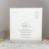 A square white invitation card with delicate floral borders and printed text, displayed upright on a light surface with small yellow and pink flower petals around it.