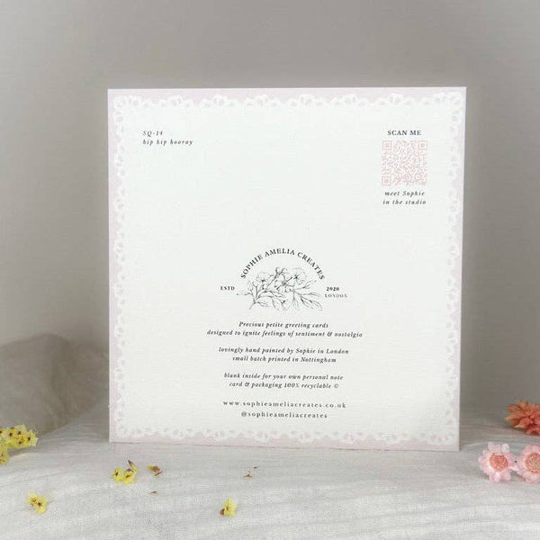 A square white invitation card with delicate floral borders and printed text, displayed upright on a light surface with small yellow and pink flower petals around it.