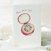 Card with a watercolor illustration of an open ring box containing a diamond ring, on a soft white background.