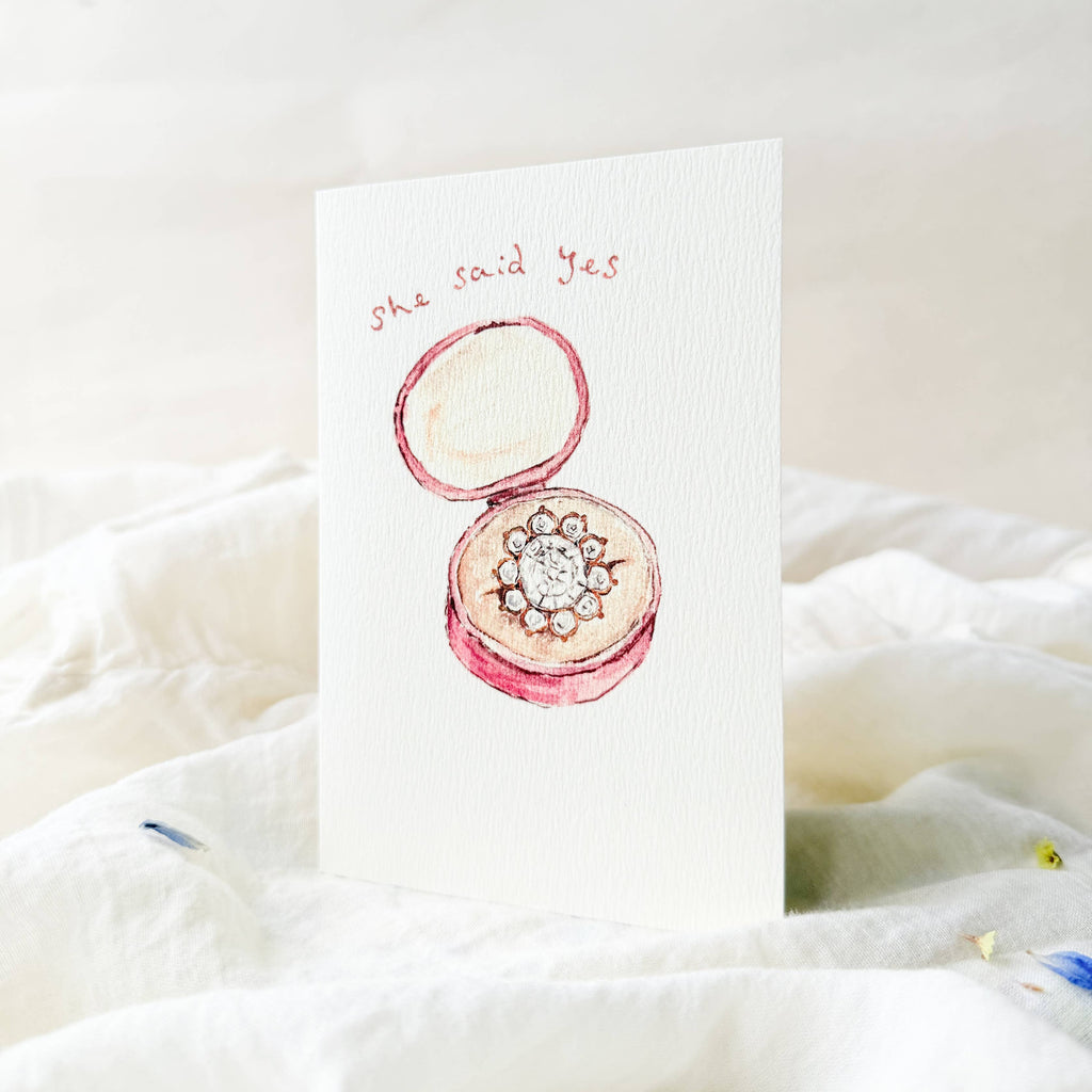 Card with a pink ring box and 'she said yes' text on a white background