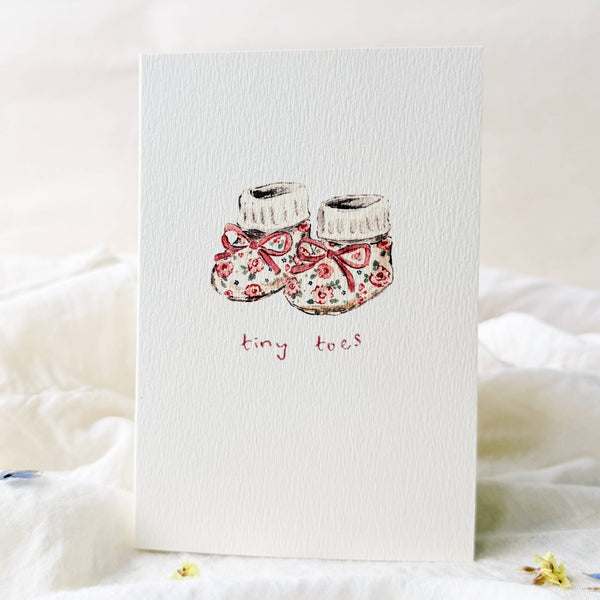 Card with illustration of baby shoes and 'tiny toes' text on a soft white background