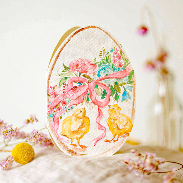 A hand-cut Easter egg card with a floral design and two chicks, presented on a surface with a decorative background.