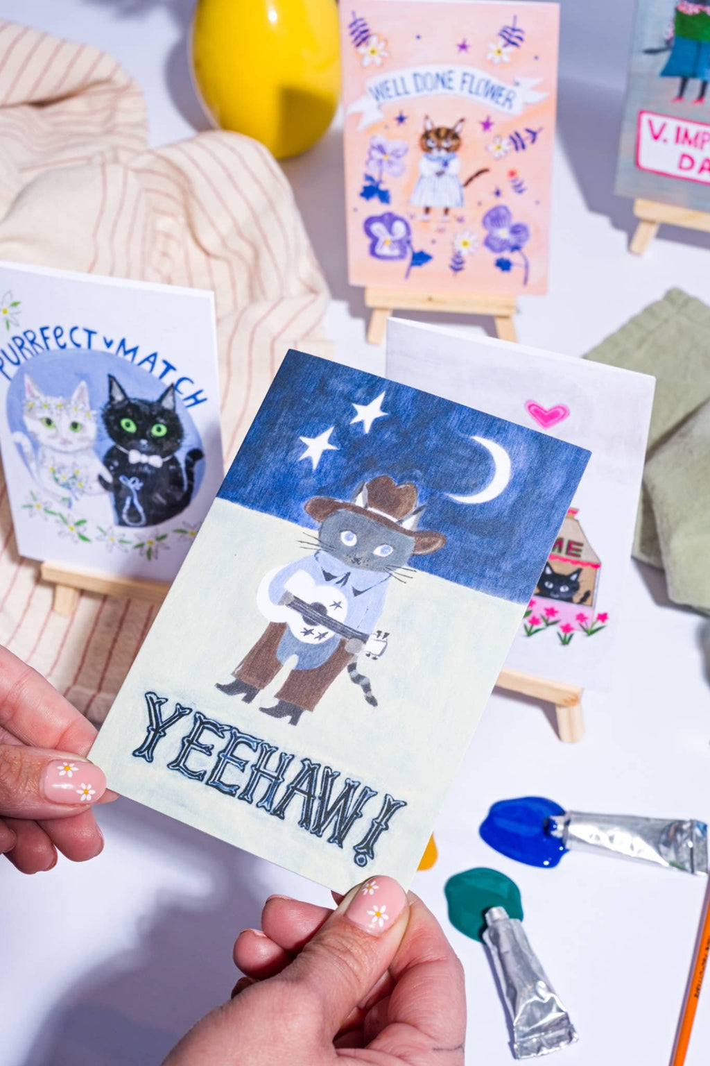 Hand holding a card with a cowboy cat illustration and 'Yeehaw!' text, surrounded by stationery items.