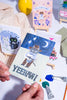 Hand holding a card with a cowboy cat illustration and 'Yeehaw!' text, surrounded by stationery items.
