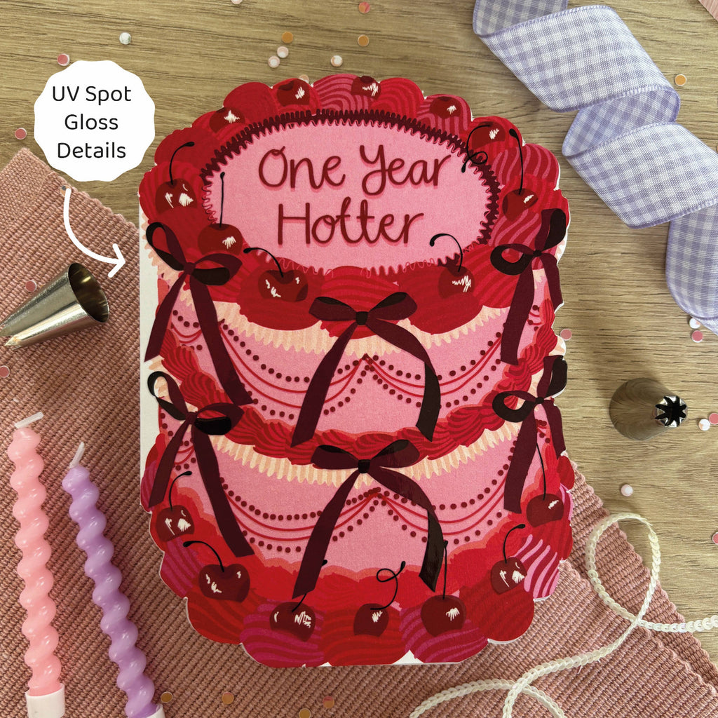 Card with red and pink design and 'One Year Hotter' text, surrounded by baking tools on a wooden surface.