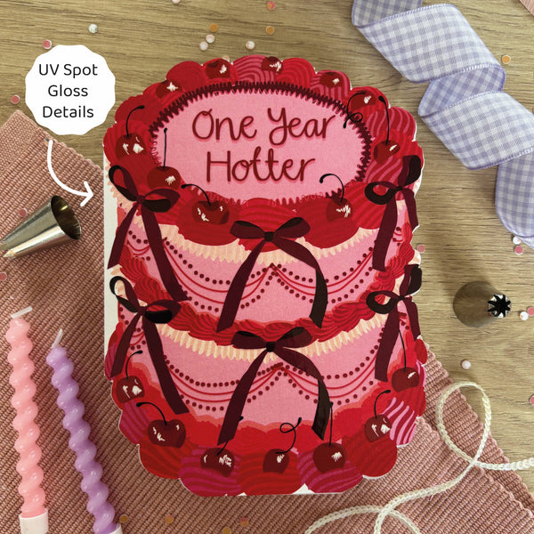 Card with red and pink design and 'One Year Hotter' text, surrounded by baking tools on a wooden surface.