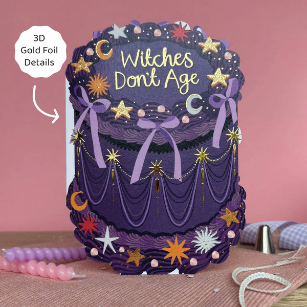 Decorative cake with 'Witches Don't Age' text and gold foil details on a pink background