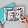 Hand holding a birthday card with decorative border and 'Happy Birthday' text, against a gray background.
