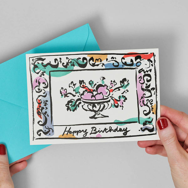 Hand holding a birthday card with decorative border and 'Happy Birthday' text, against a gray background.