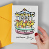 Hand holding a card with a colorful carousel illustration and 'Welcome Baby' text, next to a yellow envelope.