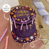 Decorative purple cake-shaped card with 'Witches Don't Age' text, gold foil details, and festive decorations on a wooden surface.