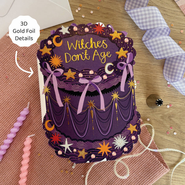 Decorative purple cake-shaped card with 'Witches Don't Age' text, gold foil details, and festive decorations on a wooden surface.