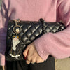 Person holding a black quilted handbag with a cat charm, wearing a pink sweater.