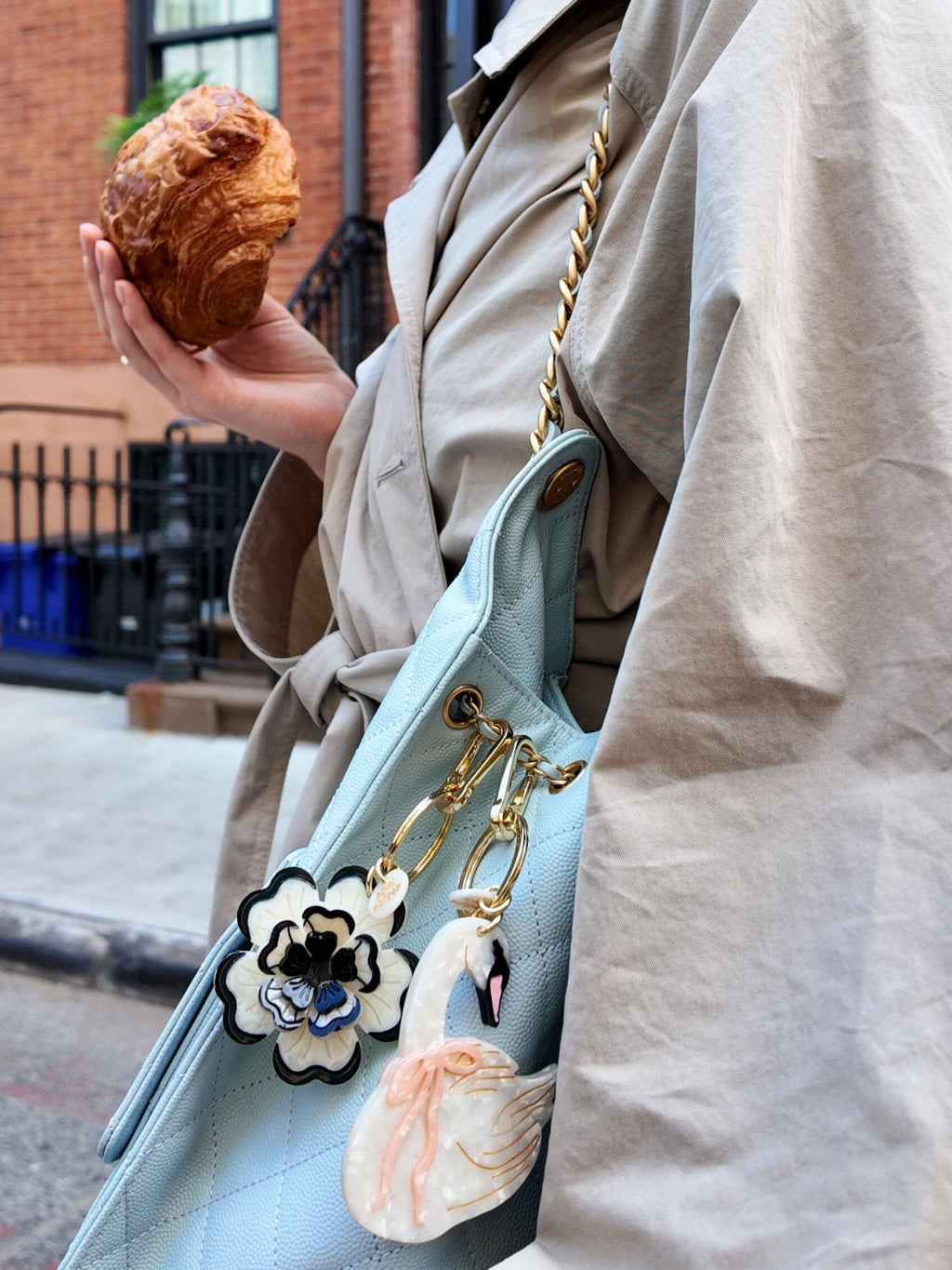 Person holding a light blue handbag with decorative charms, wearing a beige coat outdoors.