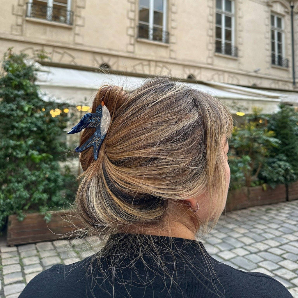 Person with styled hair in an outdoor setting with a building and plants in the background