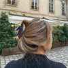 Person with styled hair in an outdoor setting with a building and plants in the background