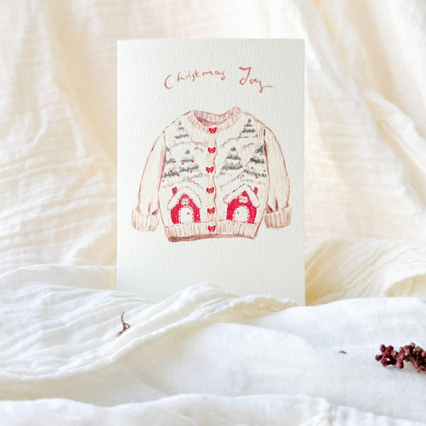 A vertical greeting card featuring a hand-drawn illustration of a Christmas sweater with pine trees and red houses, placed on a soft, draped cream-colored fabric background.