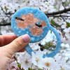 Hand holding a blue object with cherry blossom design against a cherry blossom tree background