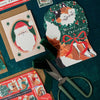 Christmas-themed cards with a cat illustration and Santa Claus, scissors on a green surface.