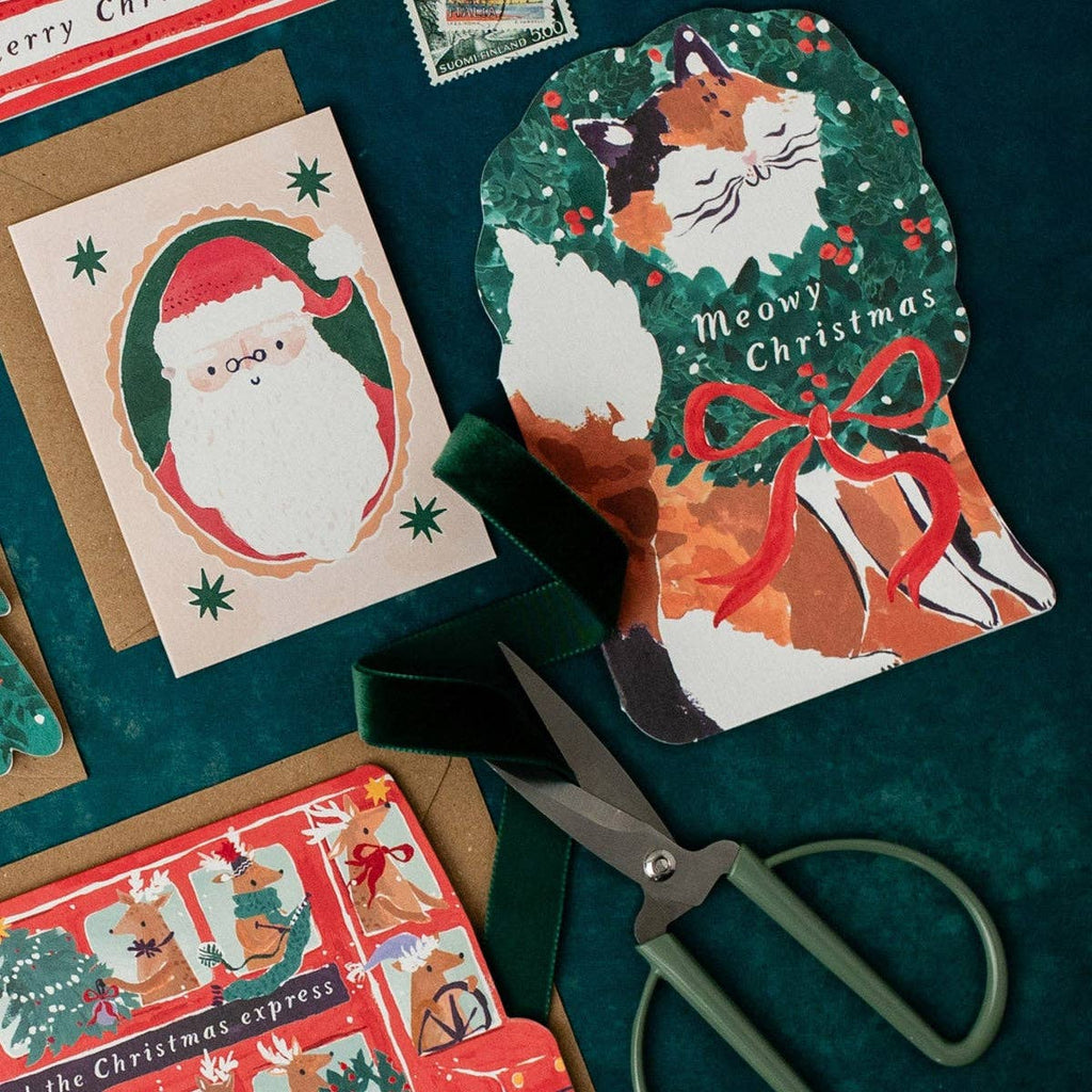 Christmas-themed cards with a cat illustration and Santa Claus, scissors on a green surface.