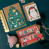 Three Christmas-themed greeting cards on a dark green surface with stationery items.