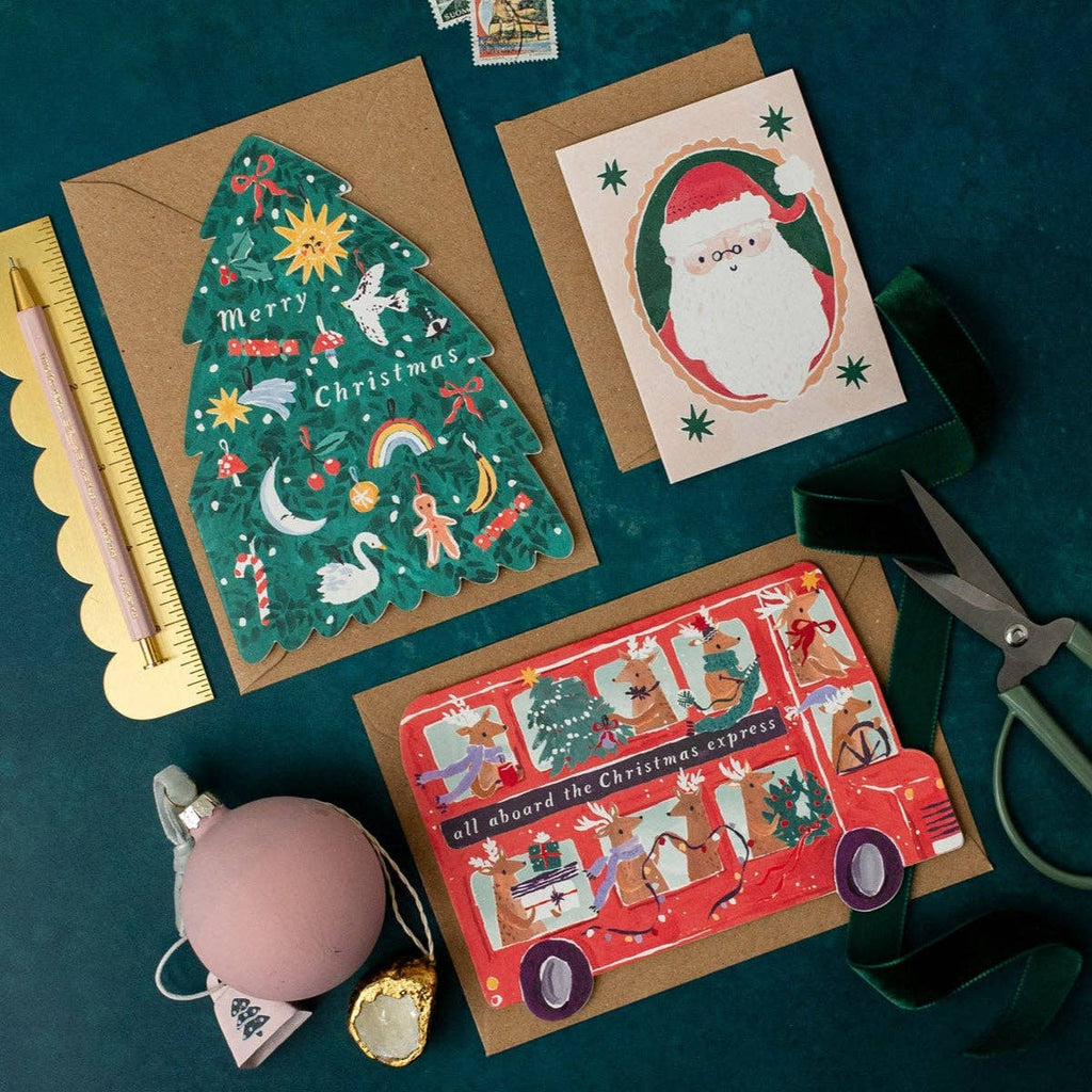 Three Christmas-themed greeting cards on a dark green surface with stationery items.
