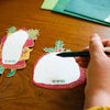 Hand using a pen on a white card with 'Last End Press' branding on a wooden surface.
