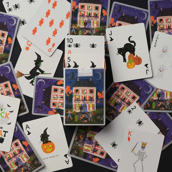 A flat-lay arrangement of Halloween-themed playing cards and a matching card deck box on a dark background. The cards feature whimsical illustrations of haunted houses, witches, skeletons, pumpkins, bats, and spiders in bright colors.