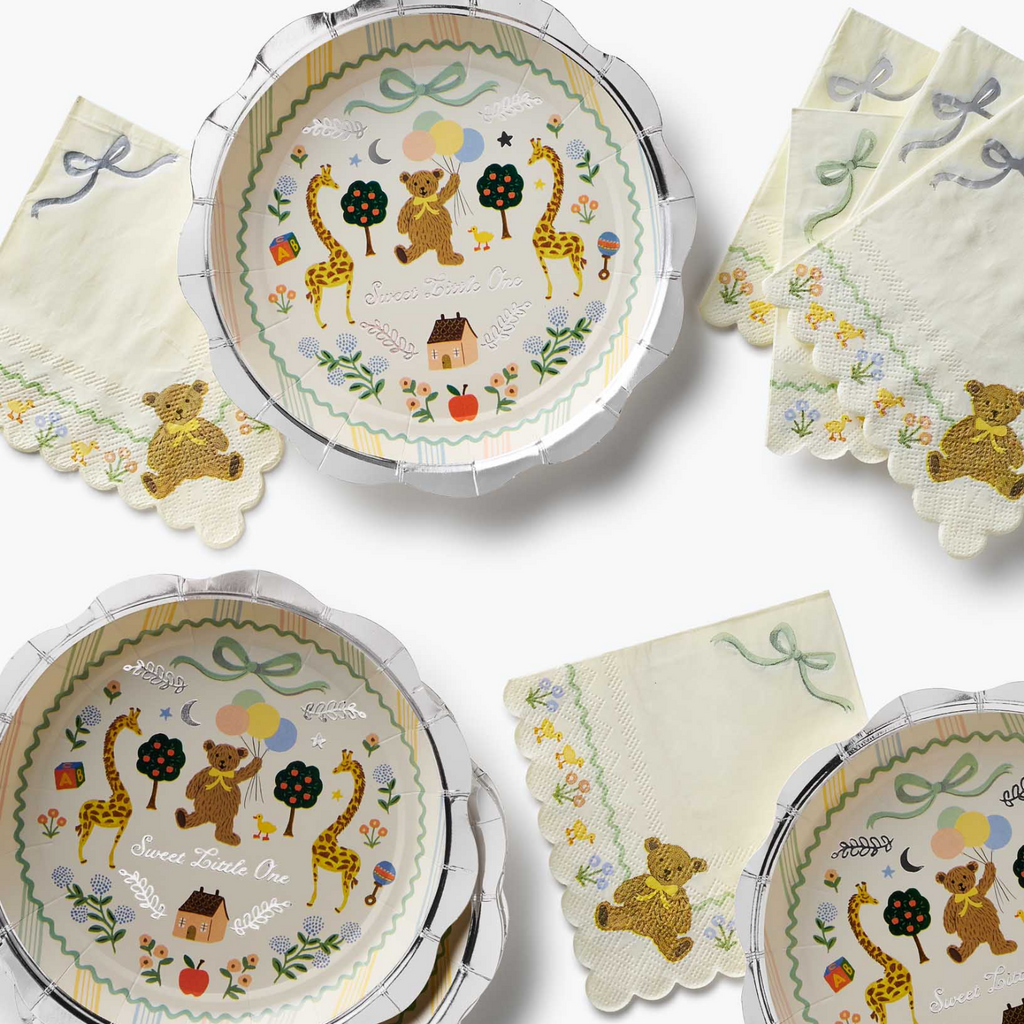 A coordinated baby shower tableware set featuring scalloped paper plates and napkins with a whimsical safari-themed embroidery design. The set includes a teddy bear, giraffes, trees, balloons, and a small house, with 'Sweethearts One' embroidered on the napkins.