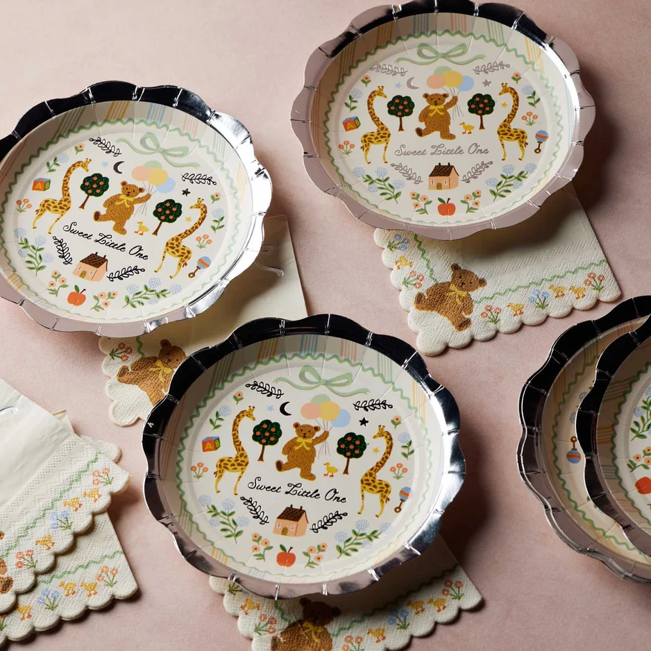 A set of scalloped paper plates and matching napkins with a whimsical woodland theme, featuring a bear, giraffes, trees, and the text 'Sweet Treats Over' on a soft pink background.