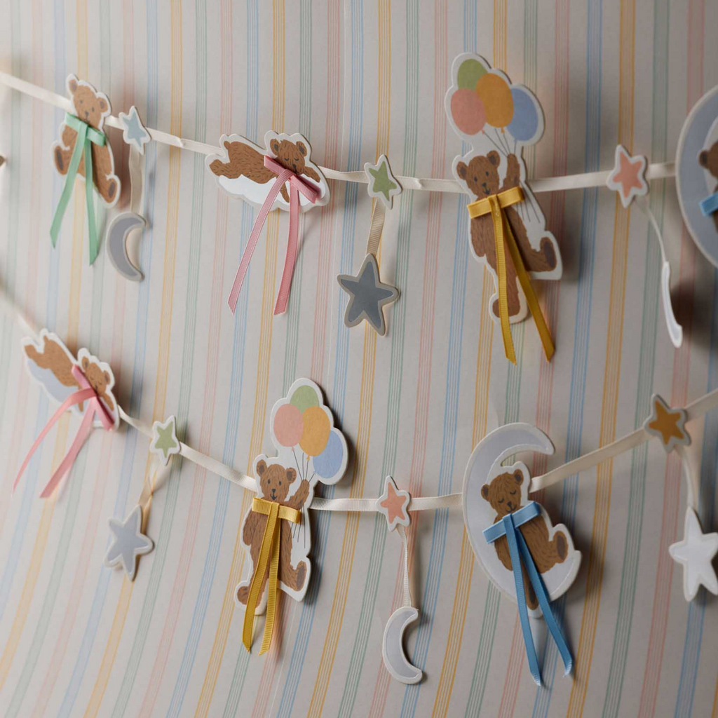 A pastel baby shower decoration garland featuring teddy bear, crescent moon, star, and balloon shapes in soft colors, hung against a striped pastel wallpaper background.