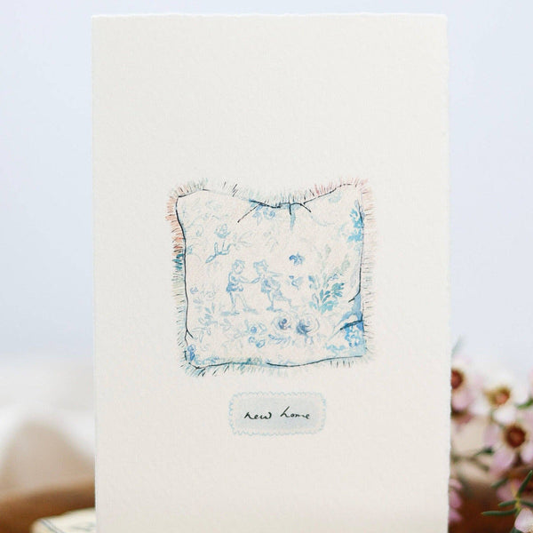 Small floral handkerchief on a card with 'New Home' text, placed on a wooden block with 'Satchel' branding.