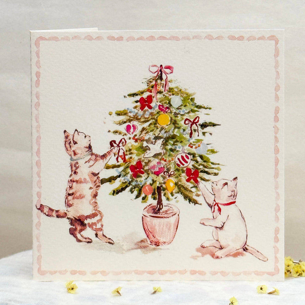 Card with illustration of two cats decorating a Christmas tree on a white surface.