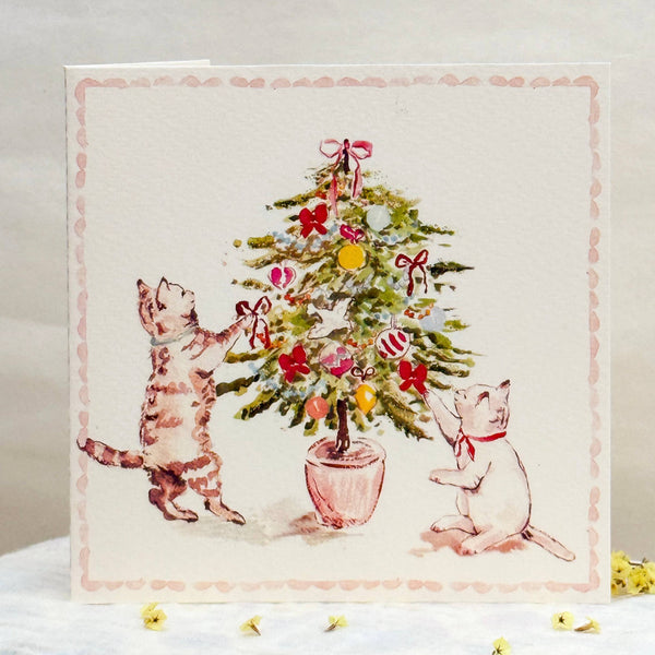 Card with illustration of two cats decorating a Christmas tree on a white surface.