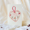 A vertical Christmas greeting card with a white textured background, featuring a red heart-shaped ornament illustration with the words 'With love of Christmas' and a small red fox. The card is photographed standing upright on draped white fabric with a few small red berries scattered around.