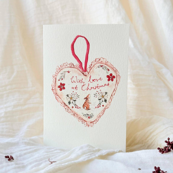 A vertical Christmas greeting card with a white textured background, featuring a red heart-shaped ornament illustration with the words 'With love of Christmas' and a small red fox. The card is photographed standing upright on draped white fabric with a few small red berries scattered around.